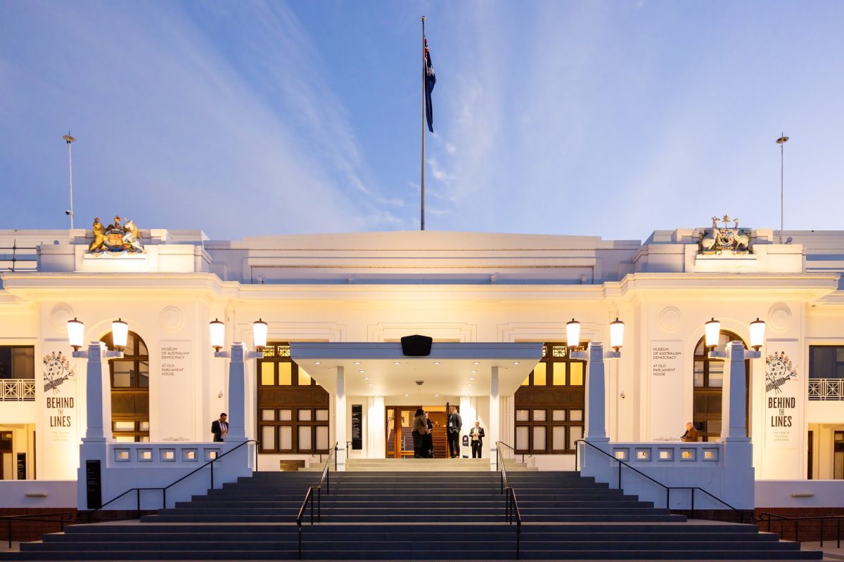 Old Parliament House lit at dusk, the Australian flag flying above the facade and guests arriving through the front entrance.