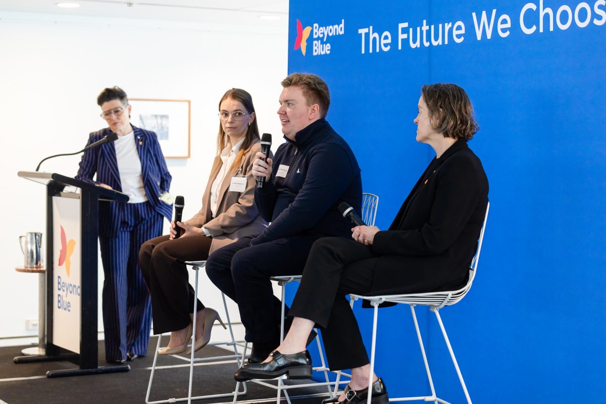• Panel on stage: The panel discussion against the Beyond Blue backdrop, three panellists seated on white wire stools with the host at the branded lectern alongside.