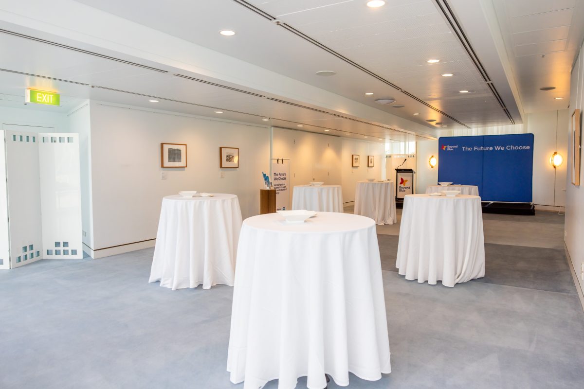 • Empty alcove wide: The alcove set for the launch, white cocktail tables running the length of the room beneath framed artworks, with the event backdrop at the far end.