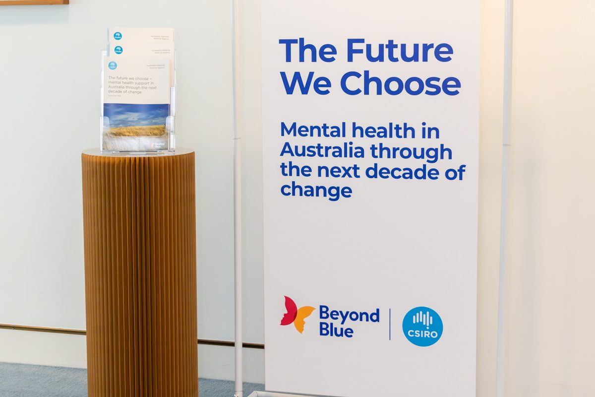 • Sign and plinth, landscape: The Future We Choose signage on a white freestanding frame beside a fluted plinth holding printed copies of the report, in the alcove before guests arrive.