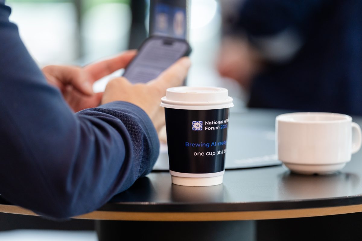 A guest resting a branded forum coffee cup on a cocktail table, the cup printed with the line about brewing AI-ready skills one cup at a time.