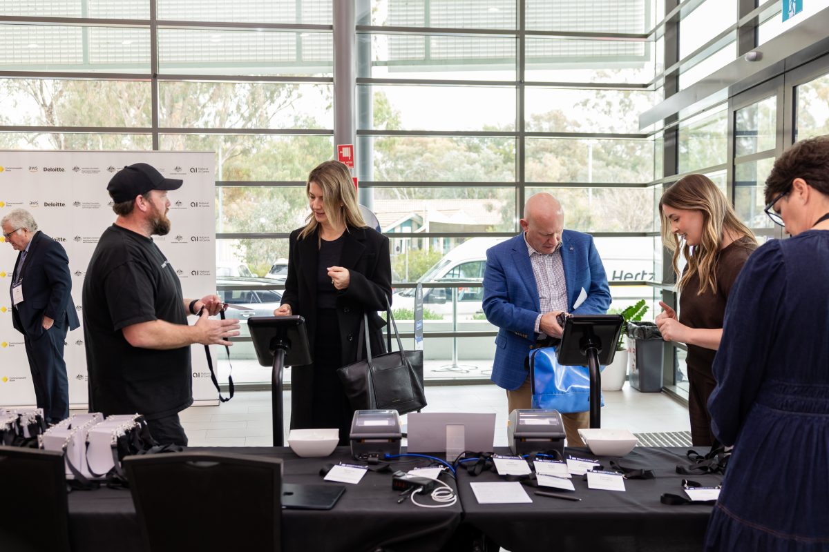 The check-in desk in use, two tablet kiosks and two label printers on the table with lanyards laid out, guests checking themselves in against the supporter media wall.
