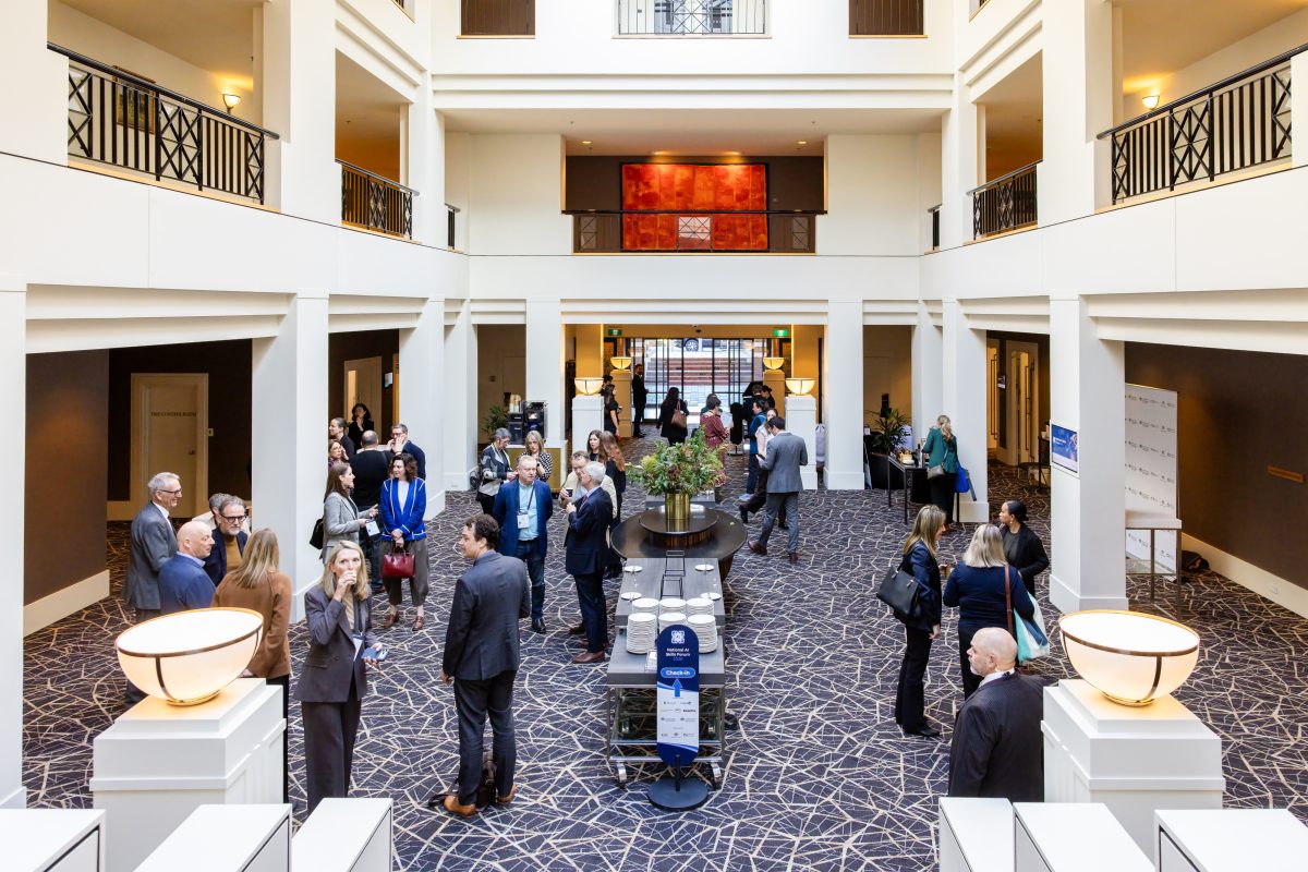 The Hyatt Hotel Canberra atrium during a break, delegates gathered around the central table with a National AI Skills Forum check-in wayfinder sign and the supporter media wall to one side.