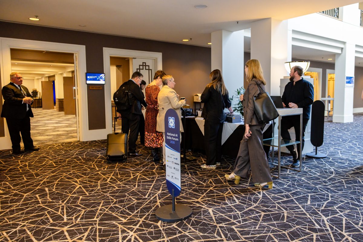 Day two check-in at the Hyatt, guests collecting badges at a compact registration table beside a forum wayfinder sign, with a digital room screen above The Gallery doors behind.