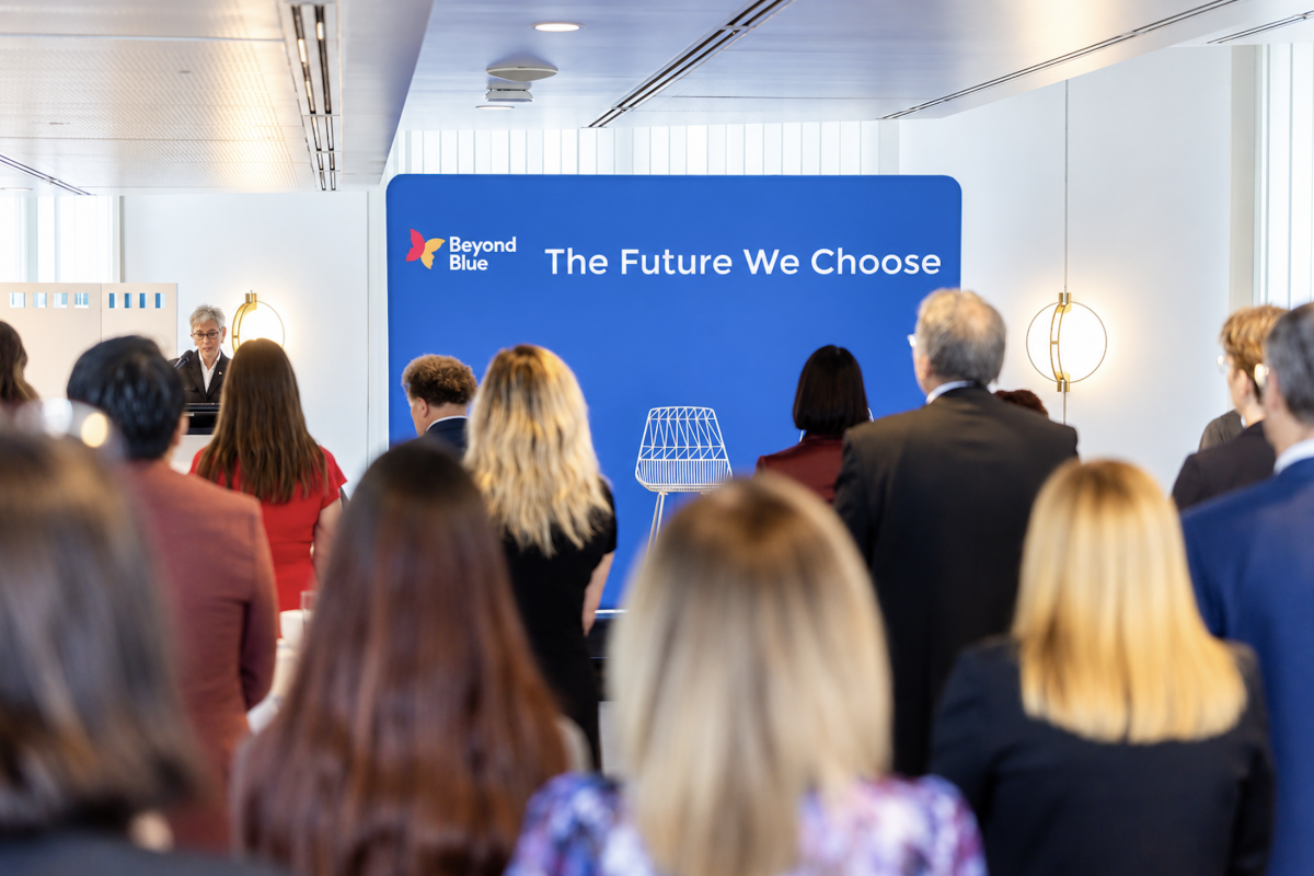 • Audience wide: The launch under way in the Dame Enid Lyons Alcove, guests standing through the room while a speaker addresses them from the branded lectern, with the event backdrop behind.