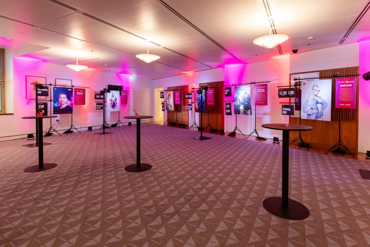 • Creator stations at the Creator Capsule launch, lit in magenta and red, with hanging panels showing a creator portrait, channel statistics and a written biography on freestanding rigs set in front of the room’s existing framed artworks.