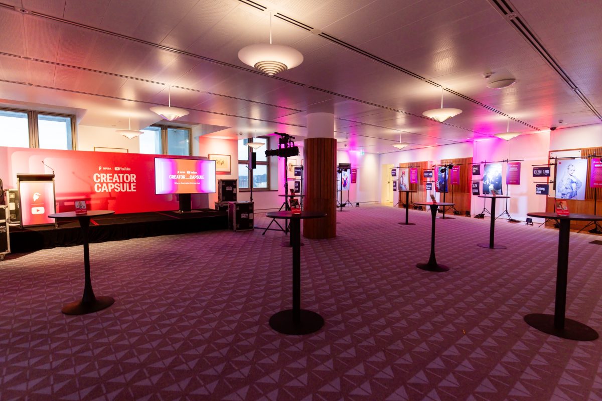 • Creator stations at the Creator Capsule launch, lit in magenta and red, with hanging panels showing a creator portrait, channel statistics and a written biography on freestanding rigs set in front of the room’s existing framed artworks.