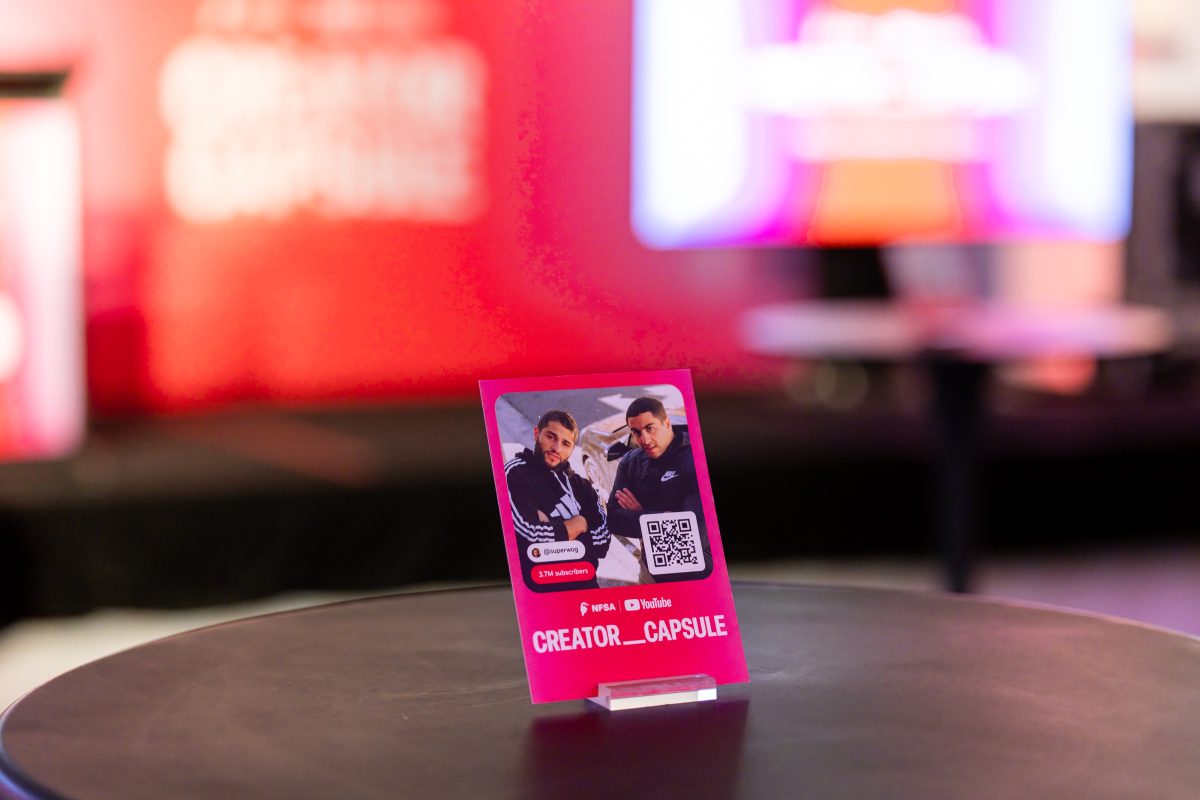 A pink Creator Capsule table card for the superwog channel, showing the creators, a QR code and subscriber count, standing on a cocktail table with the stage lit behind.