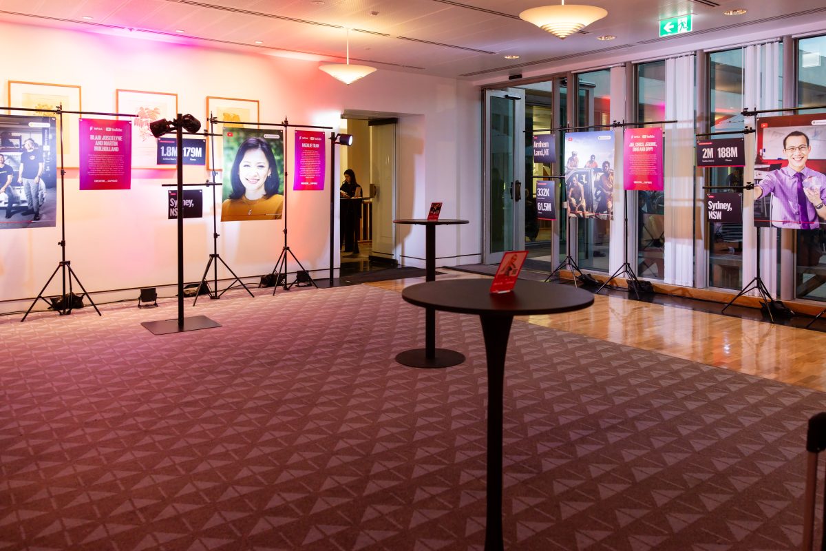 • Creator stations at the Creator Capsule launch, lit in magenta and red, with hanging panels showing a creator portrait, channel statistics and a written biography on freestanding rigs set in front of the room’s existing framed artworks.